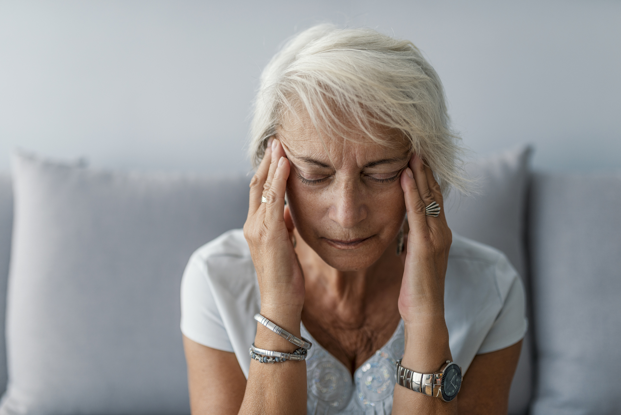 Senior woman having headache and touching her temples. Mature woman sitting on a white sofa in a home touching her head with her hands while having a headache pain and feeling unwell